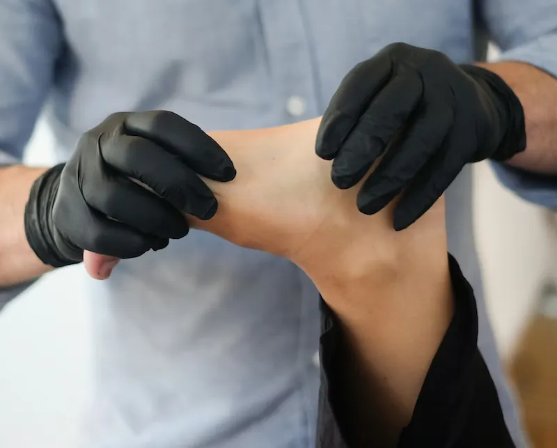 A healthcare professional in black gloves performing a physical exam on a patient's foot to identify nerve damage.