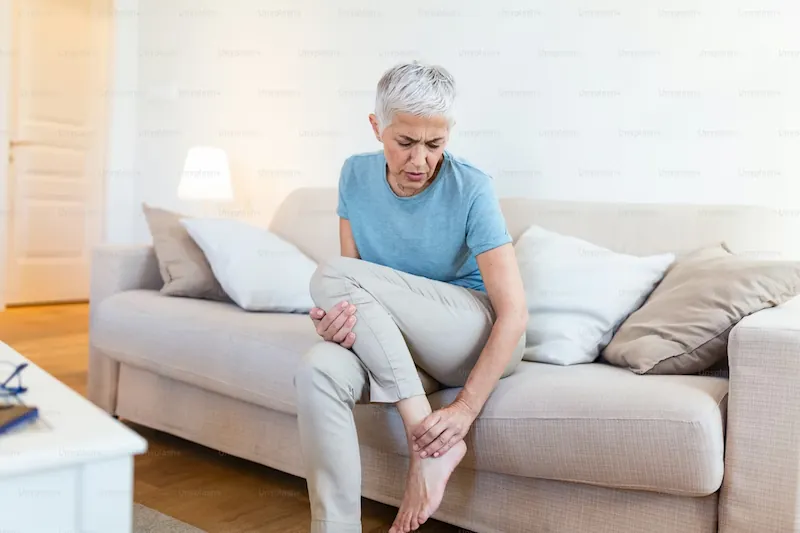An elderly woman sitting on a sofa and holding her painful foot, illustrating how peripheral neuropathy affects daily life and mobility.