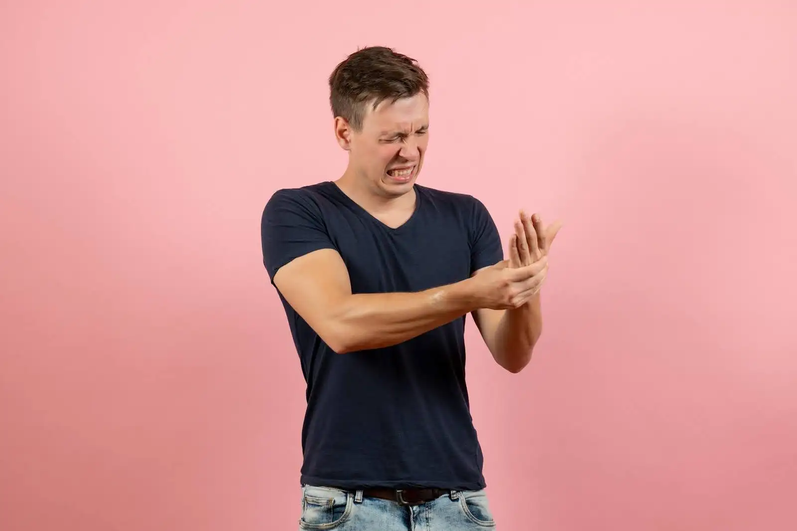 A young man wearing a navy blue t-shirt and jeans grimaces in pain while holding his wrist and hand.