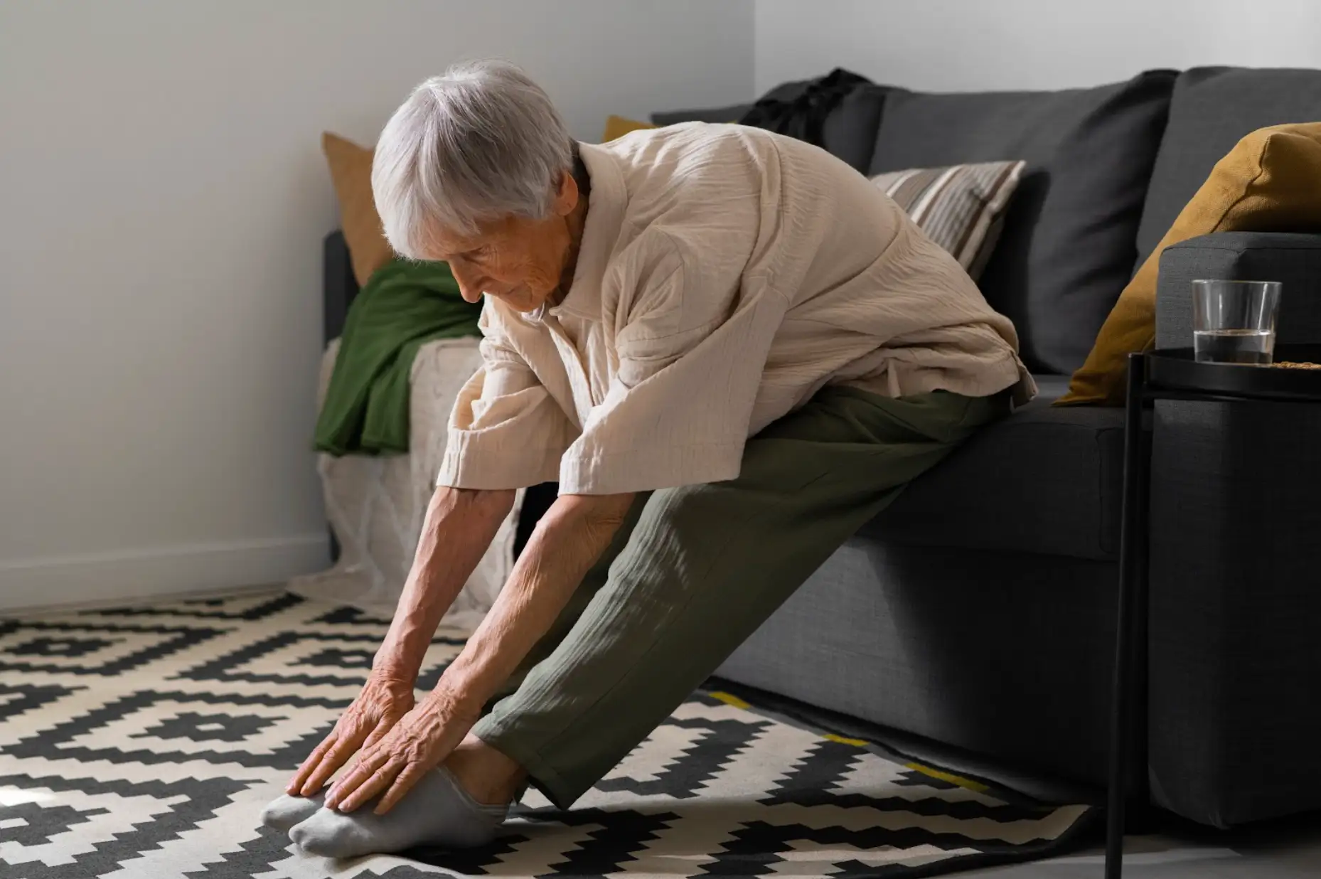 An elderly person sitting on a couch, stretching forward to touch their toes.